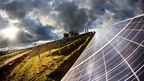 Eine Solaranlage auf der grünen Wiese. Im Hintergrund rauchen die Schlote eines Kraftwerks. 