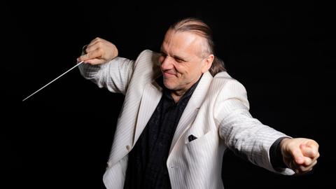 Ein Dirigent in einem hellen Jacket streckt einerseits eine Faust von sich, auf der anderen lenkt er seinen Dirigentenstab. Ein Dirigent in einem hellen Jacket streckt einerseits eine Faust von sich, auf der anderen lenkt er seinen Dirigentenstab.