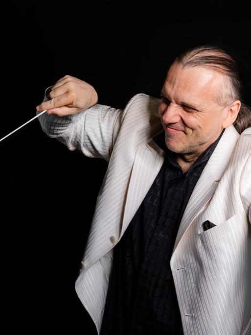 Ein Dirigent in einem hellen Jacket streckt einerseits eine Faust von sich, auf der anderen lenkt er seinen Dirigentenstab.