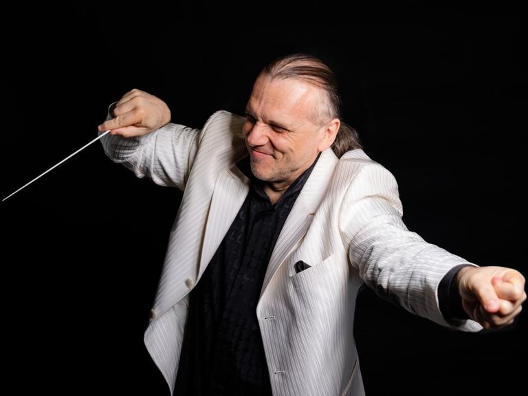 Ein Dirigent in einem hellen Jacket streckt einerseits eine Faust von sich, auf der anderen lenkt er seinen Dirigentenstab.