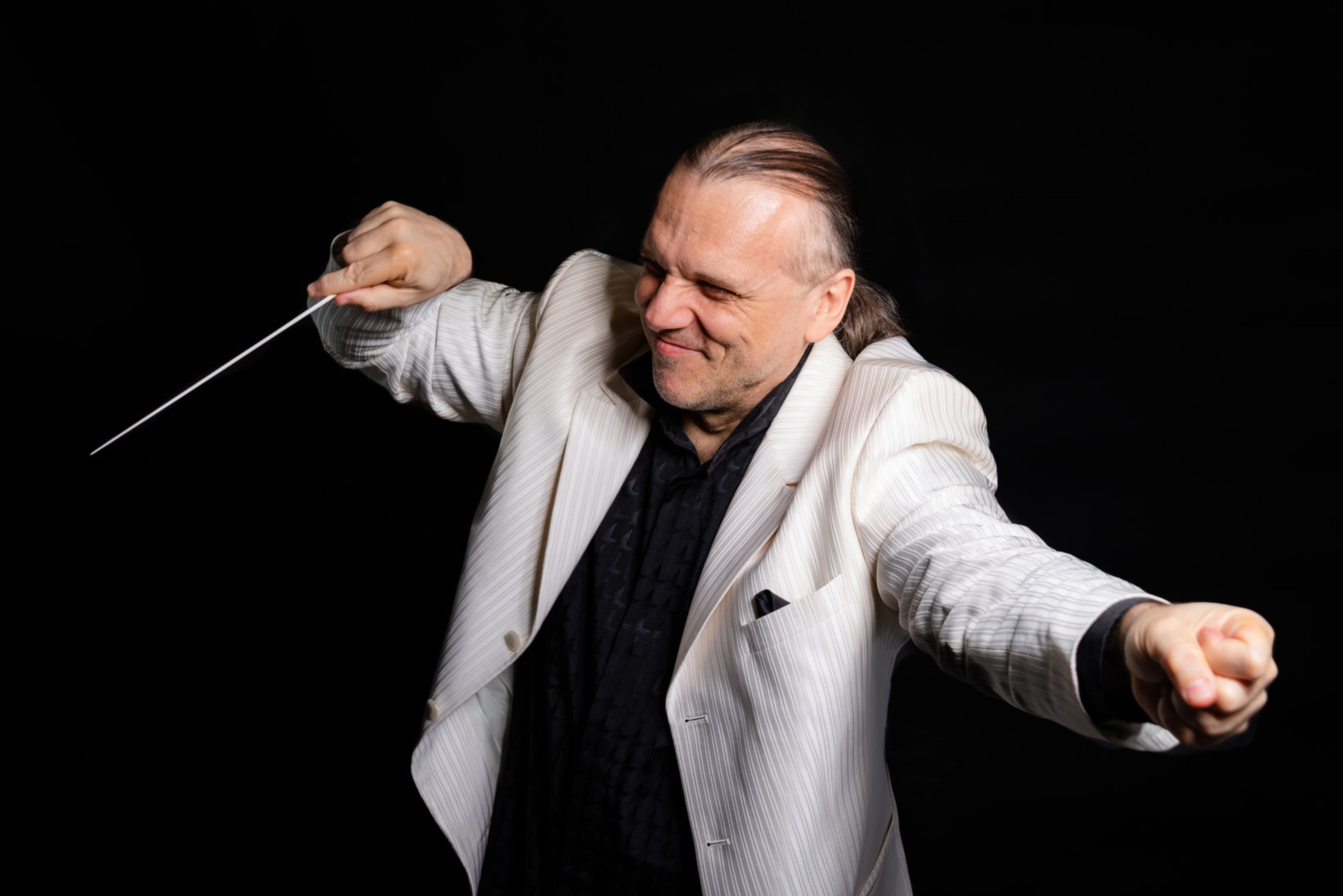 Ein Dirigent in einem hellen Jacket streckt einerseits eine Faust von sich, auf der anderen lenkt er seinen Dirigentenstab.