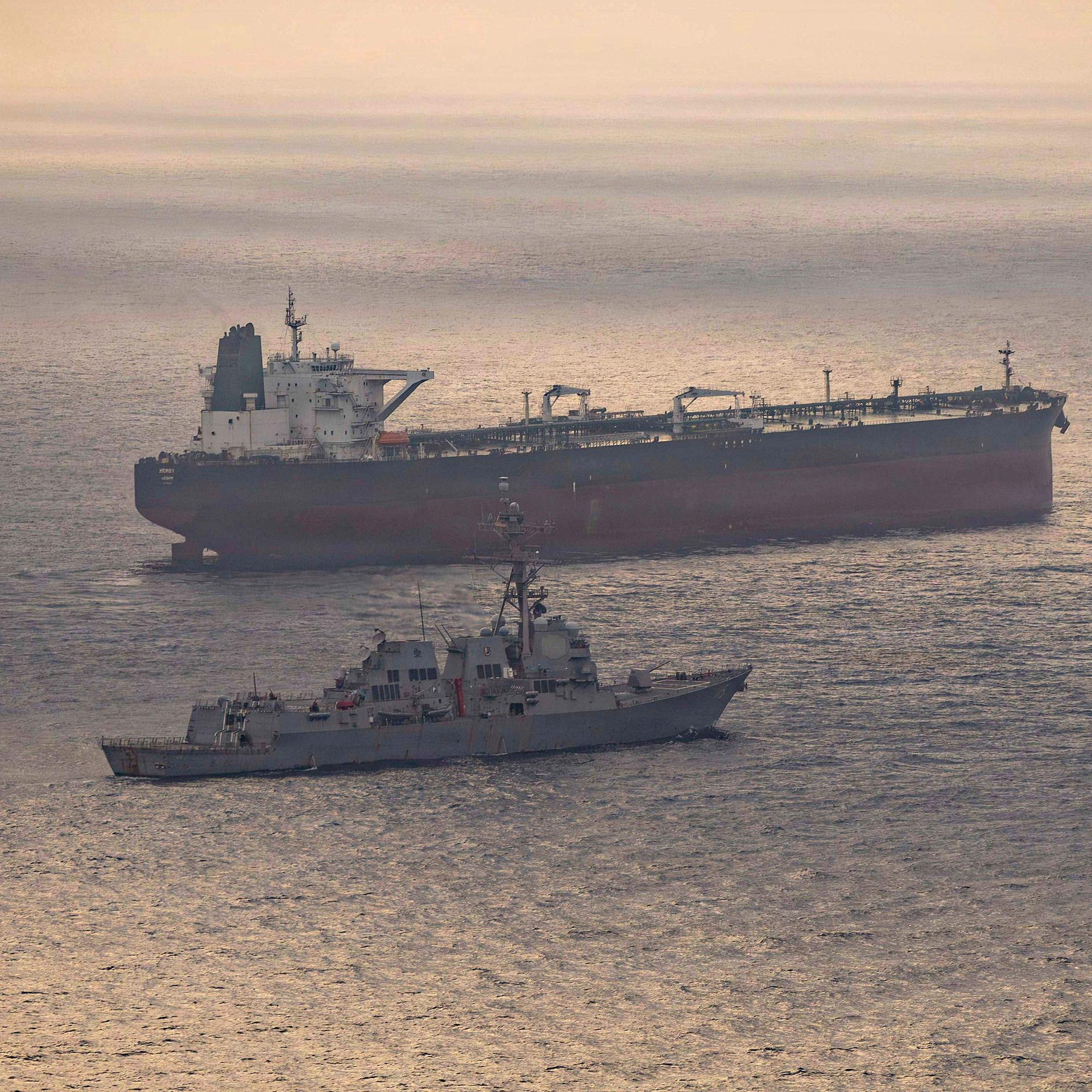 Ein US-Kriegsschiff und ein iranischer Tanker Anfang Mai in der Straße von Hormus. Auf dem Wasser spiegelt sich die untergehende Sonne.