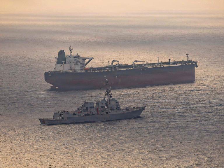 Ein US-Kriegsschiff und ein iranischer Tanker Anfang Mai in der Straße von Hormus. Auf dem Wasser spiegelt sich die untergehende Sonne.