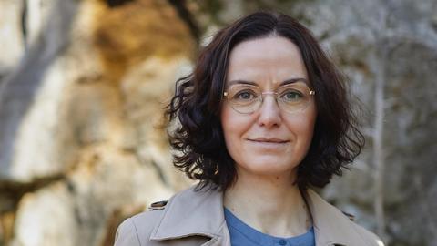 Porträt der Schriftstellerin Tina Pruschmann. Eine Frau mittleren Alters mit dunklen, halblangen Locken, goldener Brille steht im Trenchcoat und blauer Bluse vor einen teils sonnenbeschienenen Steinwand.