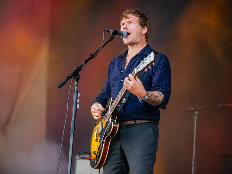 Thorsten Nagelschmidt steht mit Gitarre auf einer Konzertbühne und singt ins Mikrofon