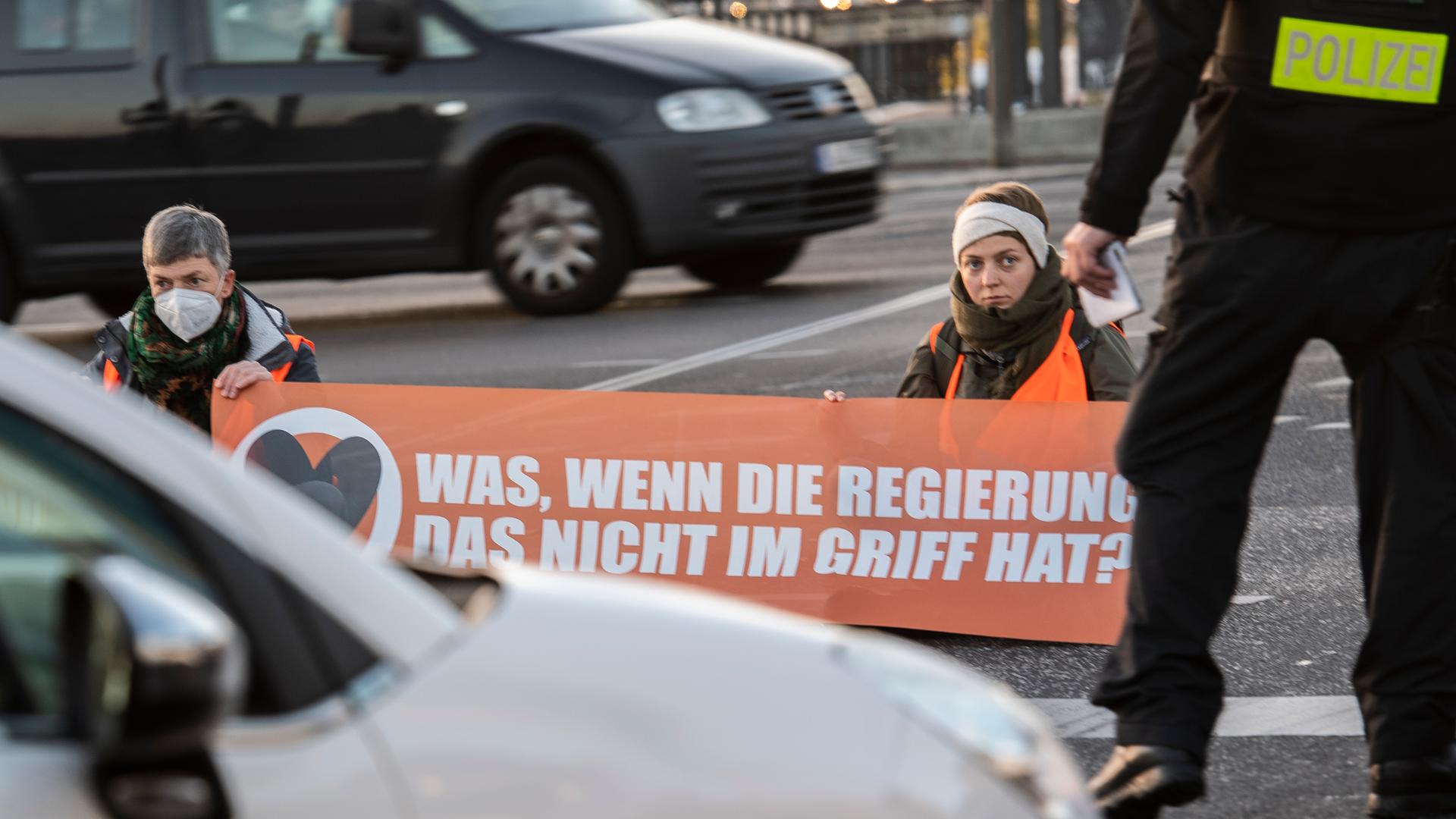 Menschen mit Transparent sitzen auf einer Straße, um sie herum Autos und Polizei.