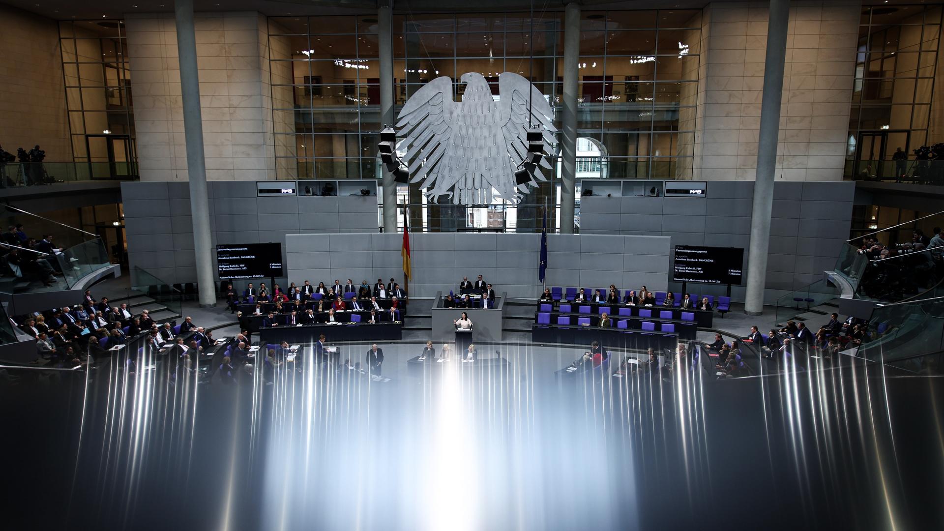 Das Bild zeigt einen weitwinkligen Blick in den Plenarsaal des Bundestags. Im Fokus steht der große Bundesadler. Darunter sind (klein) viele Abgeordnete zu sehen. 