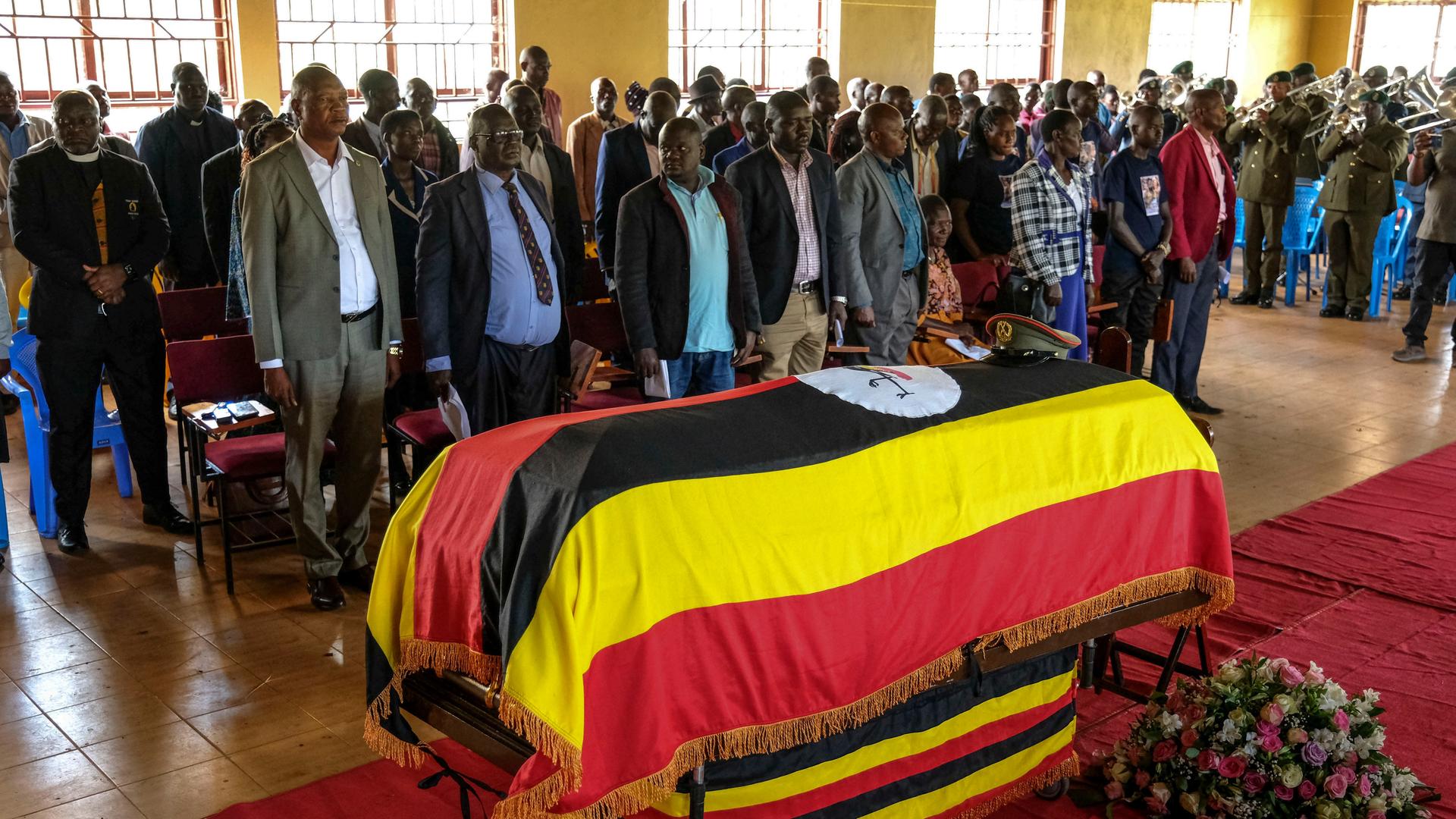 Menschen versammeln sich hinter dem Sarg der getöteten Sportlerin. Auf dem Sarg liegt die Flagge Ugandas. Menschen versammeln sich hinter dem Sarg der getöteten Sportlerin. Auf dem Sarg liegt die Flagge Ugandas.