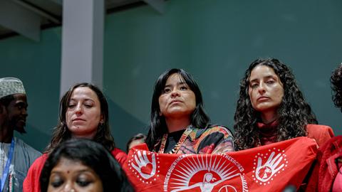 Drei Frauen stehen nebenander mit klagendem Blick. Zwei von ihnen halten einen roten Banner mit weißer Schrift hoch. Der Schriftzug beginnt mit "indigene Frauen..."