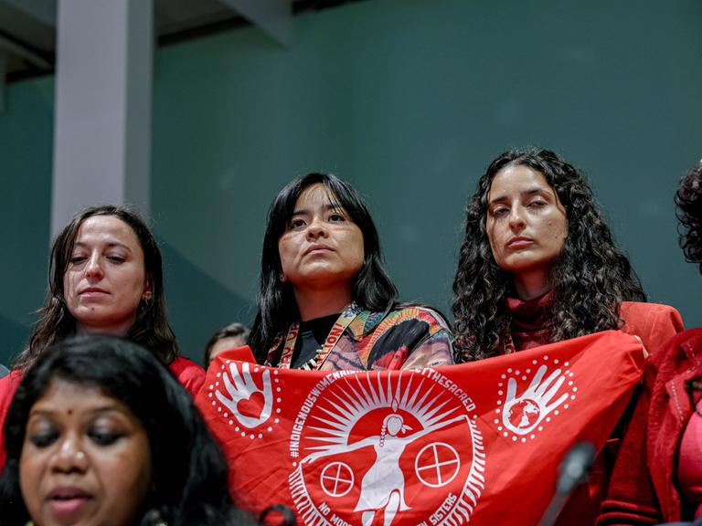 Drei Frauen stehen nebenander mit klagendem Blick. Zwei von ihnen halten einen roten Banner mit weißer Schrift hoch. Der Schriftzug beginnt mit "indigene Frauen..."