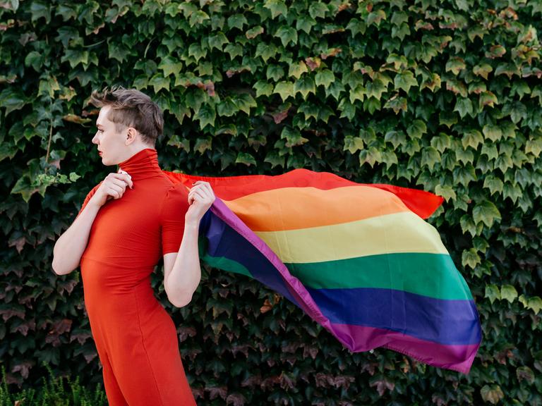 Eine nicht-binäre Person hält die Regenbogenflagge wie ein Cape an ihren Rücken.