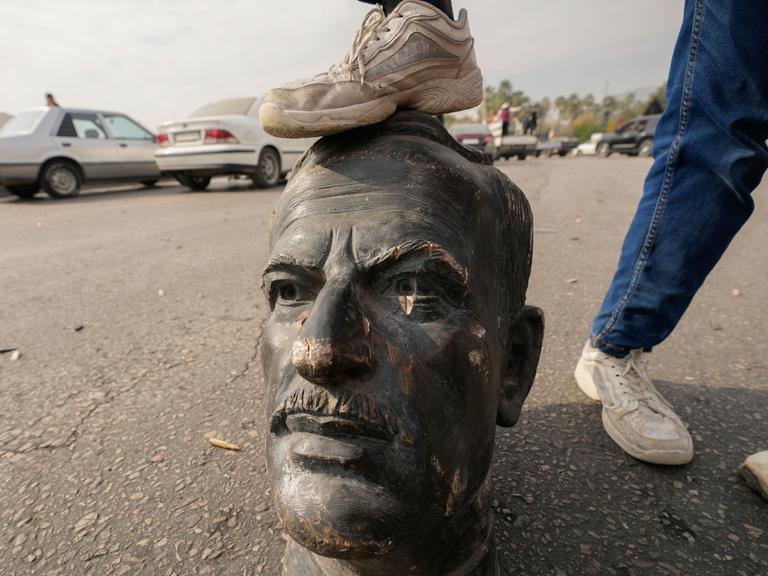 Ein Mann stellt sein Bein auf den Kopf einer zerstörten Assad-Statue auf den Straßen von Damaskus