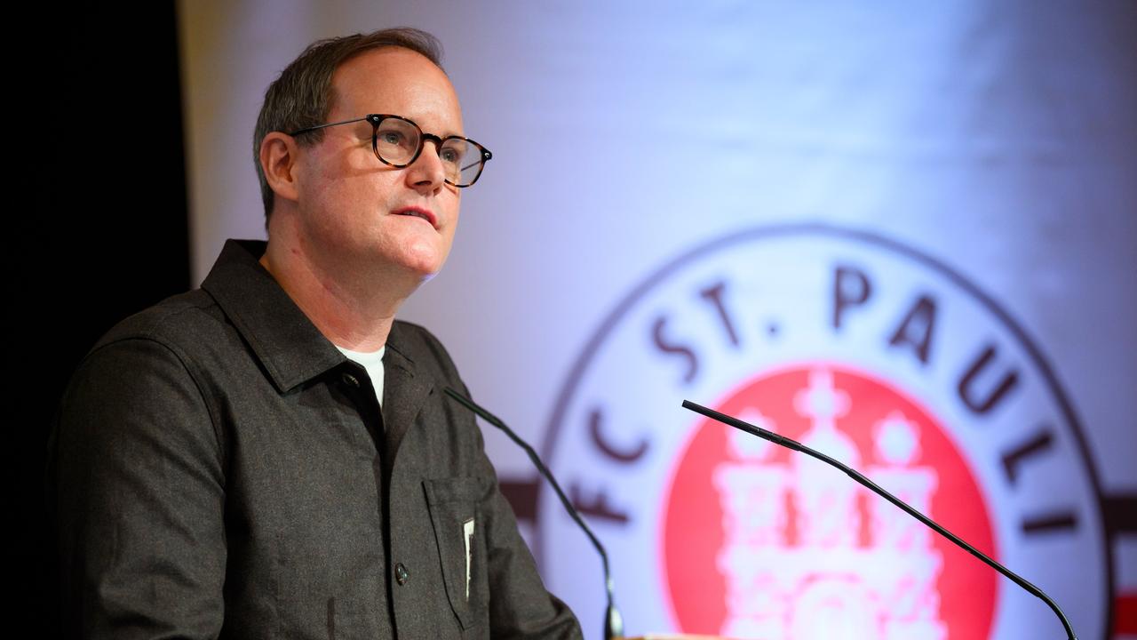 Göttlich spricht am Rednerpult in ein Mikrofon. Hinter ihm sieht man auf einer Videoleinwand das Vereinslogo.