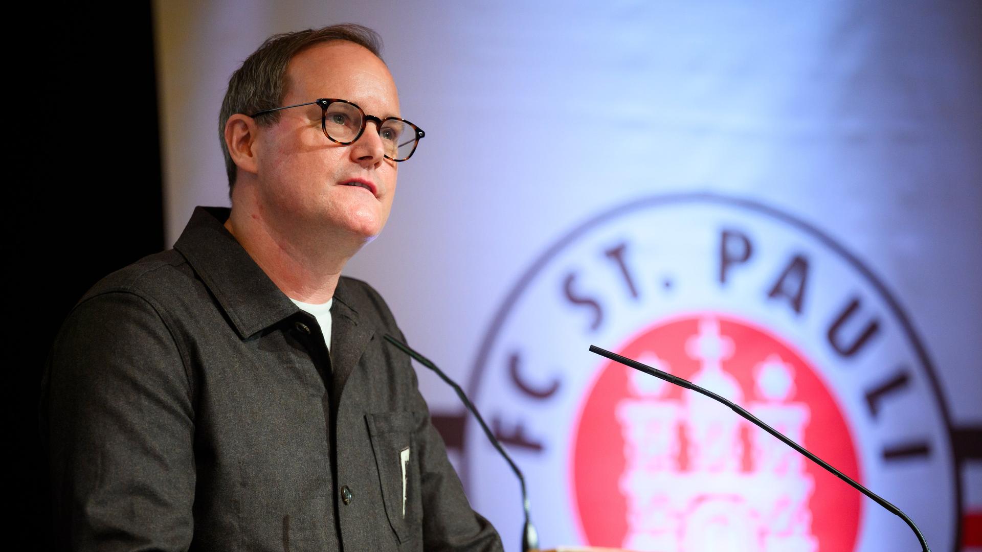 Göttlich spricht am Rednerpult in ein Mikrofon. Hinter ihm sieht man auf einer Videoleinwand das Vereinslogo. Göttlich spricht am Rednerpult in ein Mikrofon. Hinter ihm sieht man auf einer Videoleinwand das Vereinslogo.