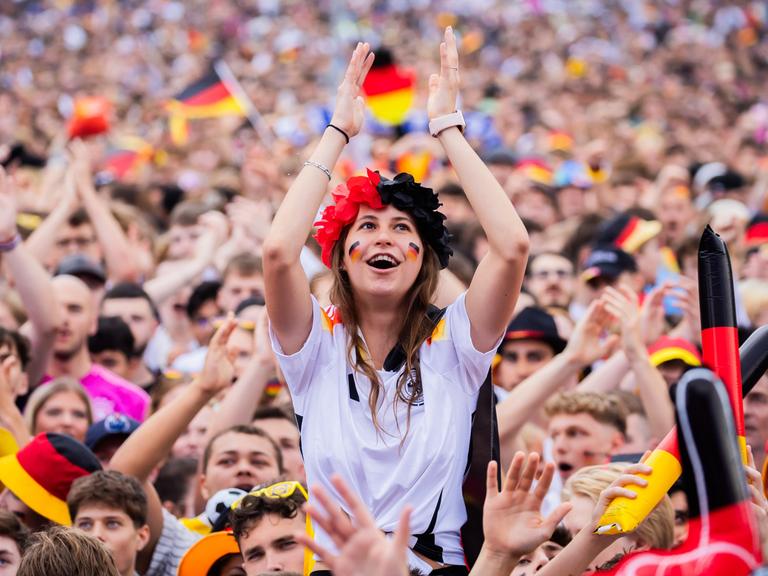 Deutschland-Fans jubeln beim Public Viewing in der Fanzone am Brandenburger Tor. Im Zentrum des Bildes ist eine Frau, die auf den Schultern einer anderen Person sitzt und jubelt.