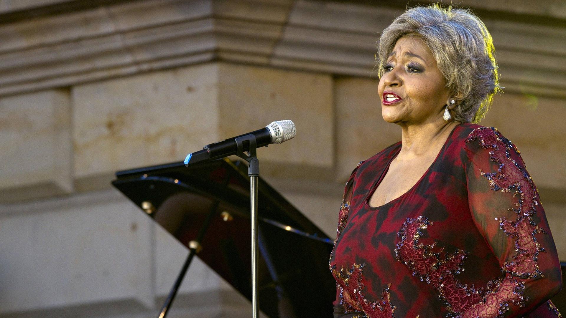 Die Opernsängerin Grace Bumbry steht in einem dunkelroten Abendkleid auf der Bühne vor einem geöffneten Flügel und singt.
