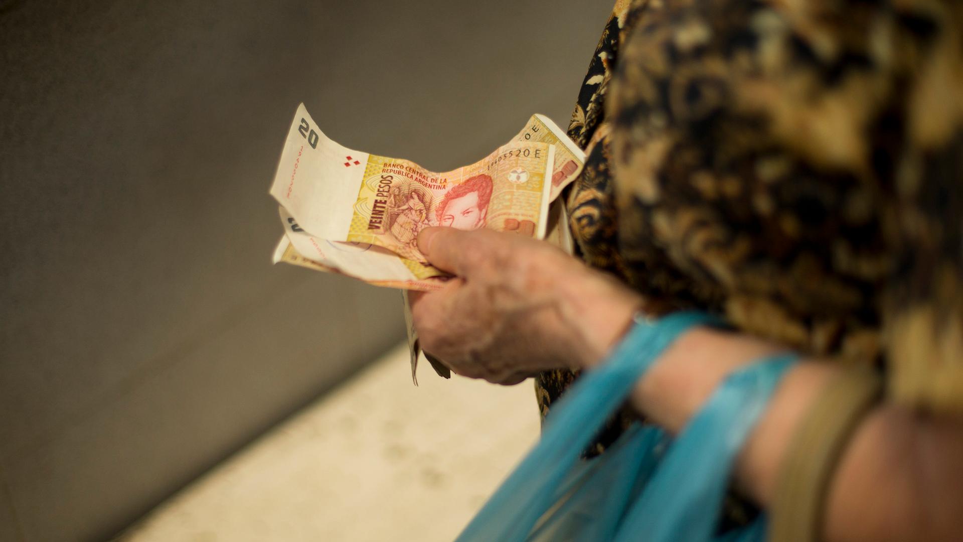 Das Foto zeigt die Hand einer älteren Frau: Sie hält darin Scheine des argentischen Pesos. Über ihrem Handgelenk hängen die Henkel einer blauen Plastiktüte. Das Foto zeigt die Hand einer älteren Frau: Sie hält darin Scheine des argentischen Pesos. Über ihrem Handgelenk hängen die Henkel einer blauen Plastiktüte.