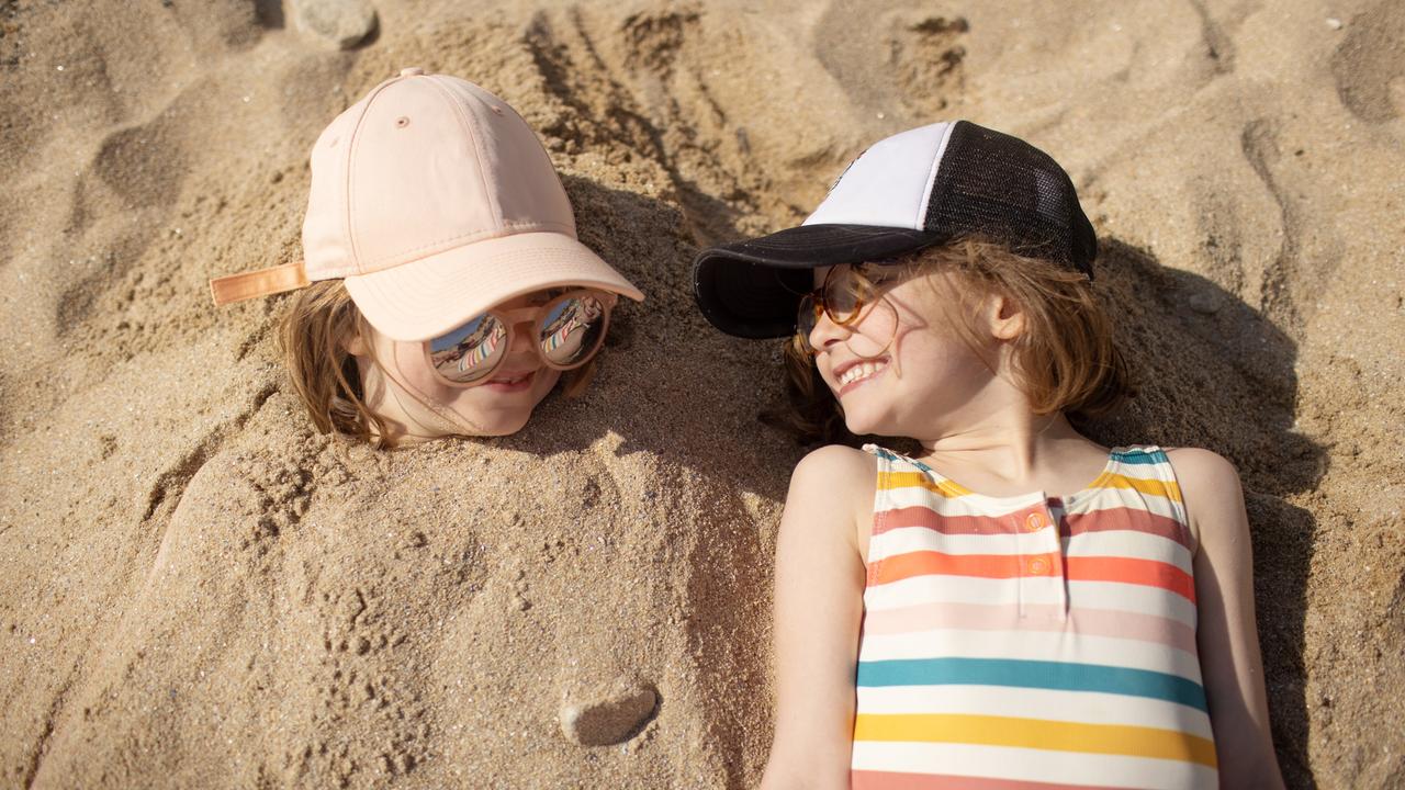 Zwei Kinder liegen nebeneinander am Strand und lachen sich an. Das linke Kind ist im Sand eingegraben. Nur der Kopf ist zu sehen.