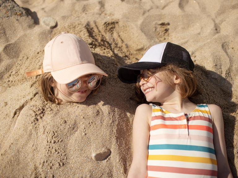 Zwei Kinder liegen nebeneinander am Strand und lachen sich an. Das linke Kind ist im Sand eingegraben. Nur der Kopf ist zu sehen.