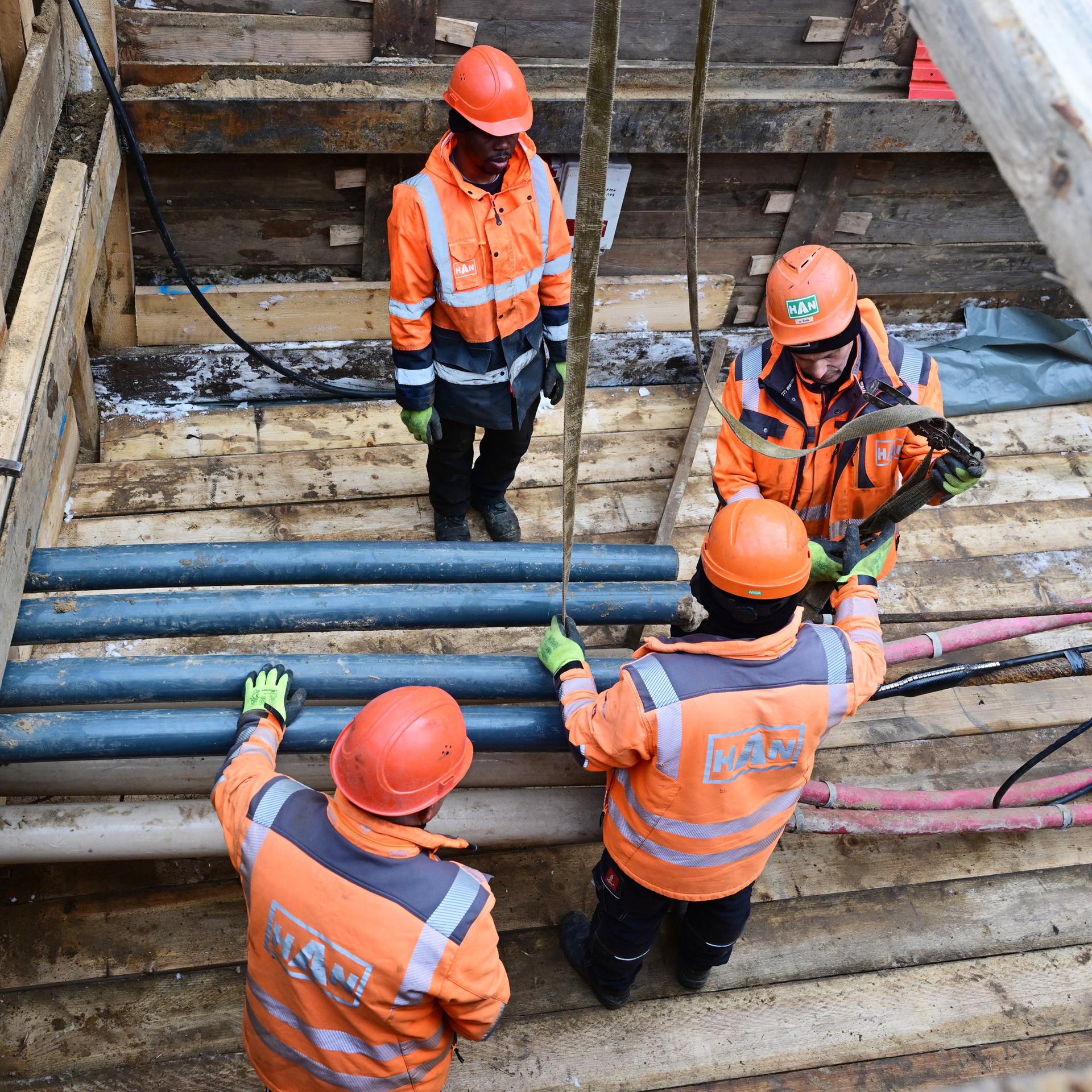 Vier Menschen mit orangefarbenen Helmen und Jacken stehen und arbeiten am 5.1.2026 in einer Baugrube mit Rohren und Kabeln.