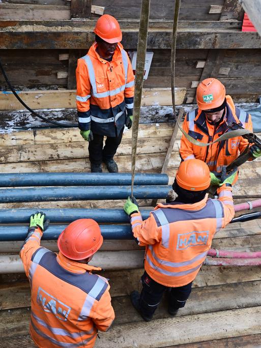 Vier Menschen mit orangefarbenen Helmen und Jacken stehen und arbeiten am 5.1.2026 in einer Baugrube mit Rohren und Kabeln.