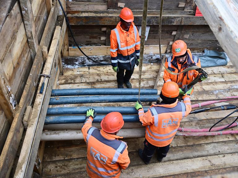 Vier Menschen mit orangefarbenen Helmen und Jacken stehen und arbeiten am 5.1.2026 in einer Baugrube mit Rohren und Kabeln.
