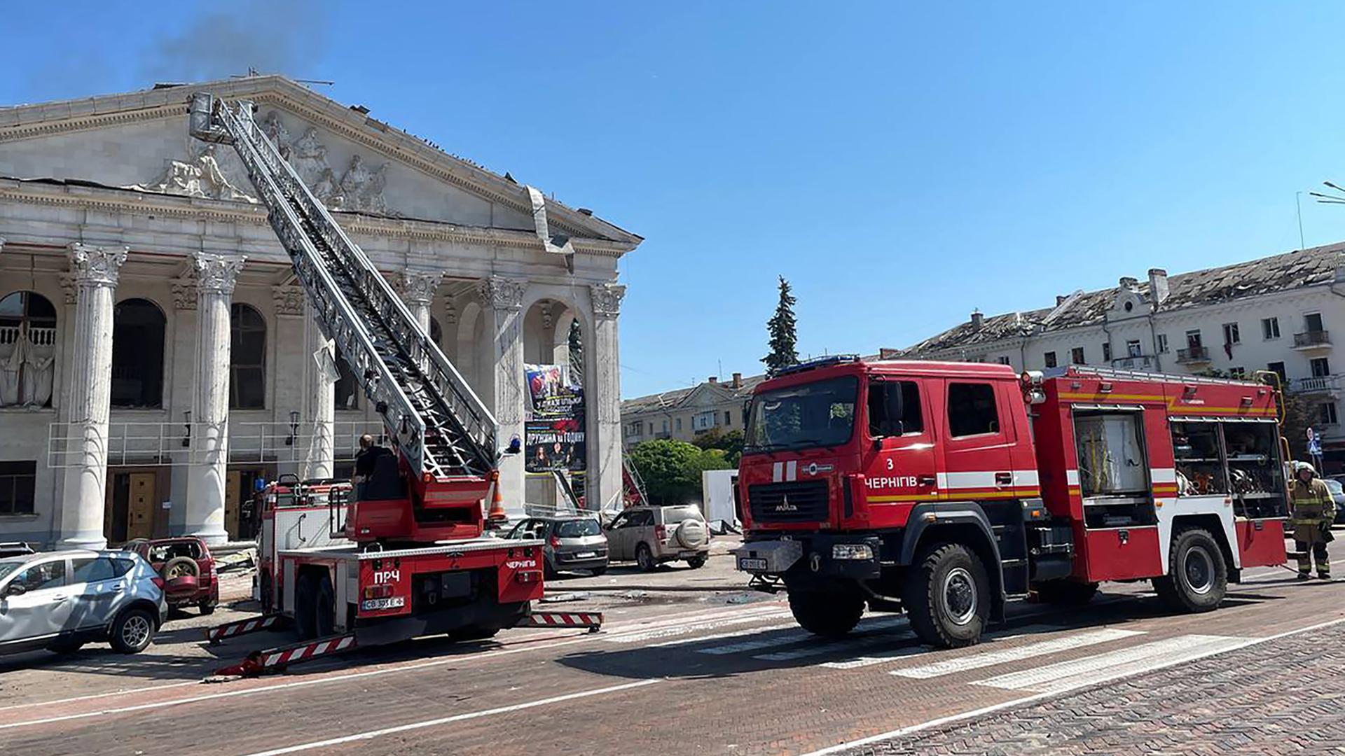 Einsatzkräfte der Feuerwehr stehen mit einer Drehleiter vor einem beschädigten Theater, aus dem Rauch steigt.