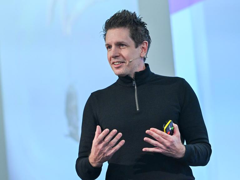 Der Medienwissenschaftler Martin Andree stehend bei einem Vortrag. Er hat kurze, dunkle Haare und einen schwarzen. hochgeschlossenen Pullover an und gestikuliert. Der Medienwissenschaftler Martin Andree stehend bei einem Vortrag. Er hat kurze, dunkle Haare und einen schwarzen. hochgeschlossenen Pullover an und gestikuliert.