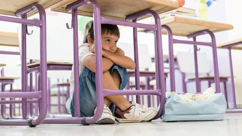 Kind sitzt unter einem Tisch in der Schule, die Knie hoch angezogen, die Arme verschränkt und das Kind darauf gestützt.