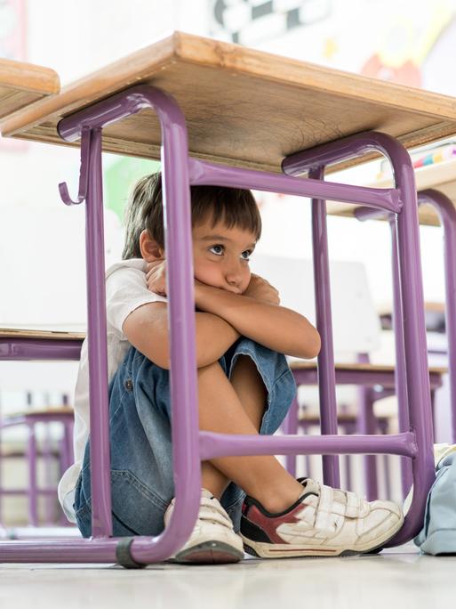 Kind sitzt unter einem Tisch in der Schule, die Knie hoch angezogen, die Arme verschränkt und das Kind darauf gestützt.