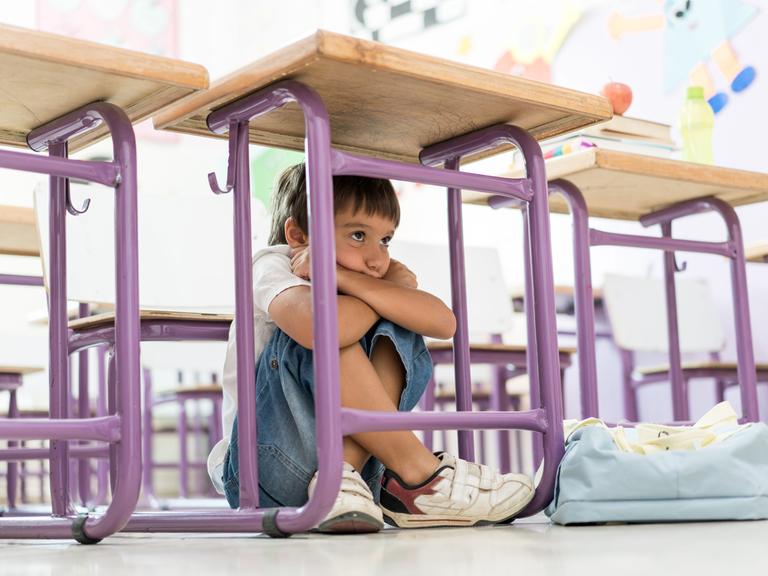 Kind sitzt unter einem Tisch in der Schule, die Knie hoch angezogen, die Arme verschränkt und das Kind darauf gestützt.