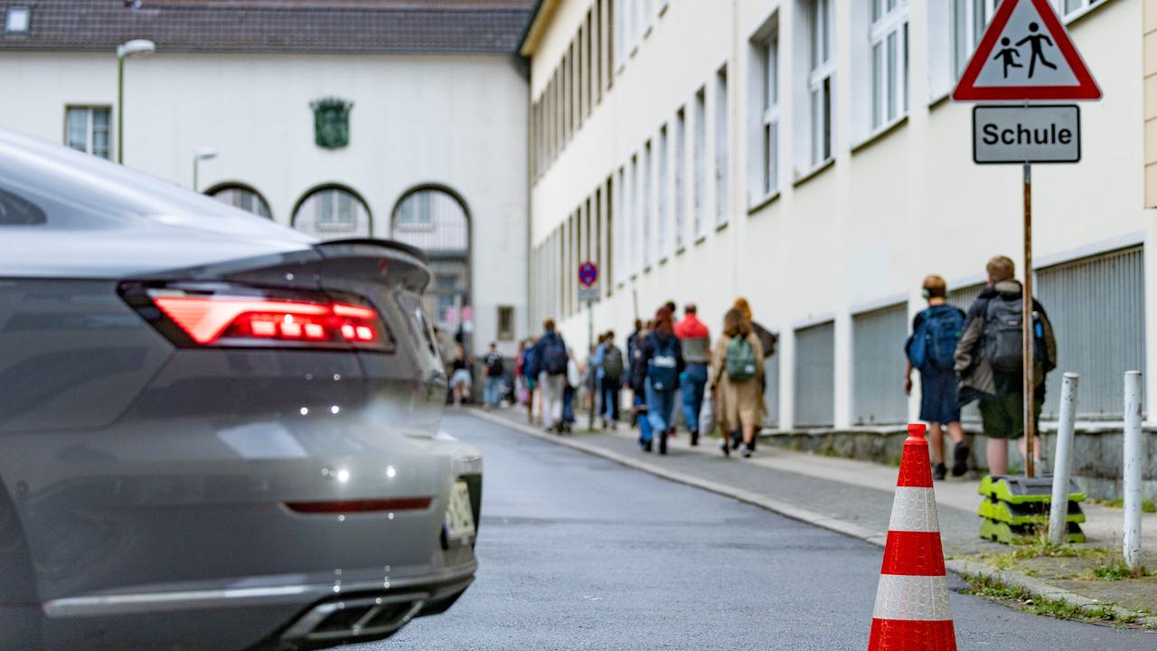 Pylonen stehen auf einer Straße vor einer Grundschule. Im Hintergrund gehen Schüler zum Schulgebäude. Im Vordergrund steht ein parkendes Auto mit rotem Bremslicht. 