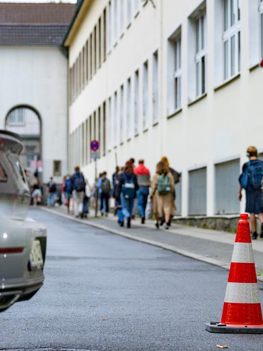 Pylonen stehen auf einer Straße vor einer Grundschule. Im Hintergrund gehen Schüler zum Schulgebäude. Im Vordergrund steht ein parkendes Auto mit rotem Bremslicht. 