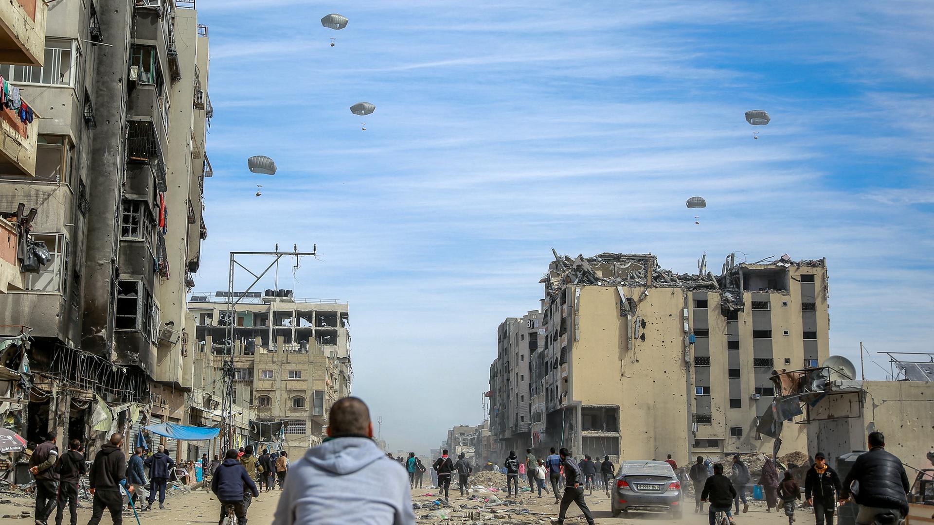 Zahlreiche Palästinenser rennen über eine vom Krieg zerstörte Straße in Richtung einiger Hilfsgüter, die an Falschirmen befestigt vom Himmel in Richtung Erde schweben.