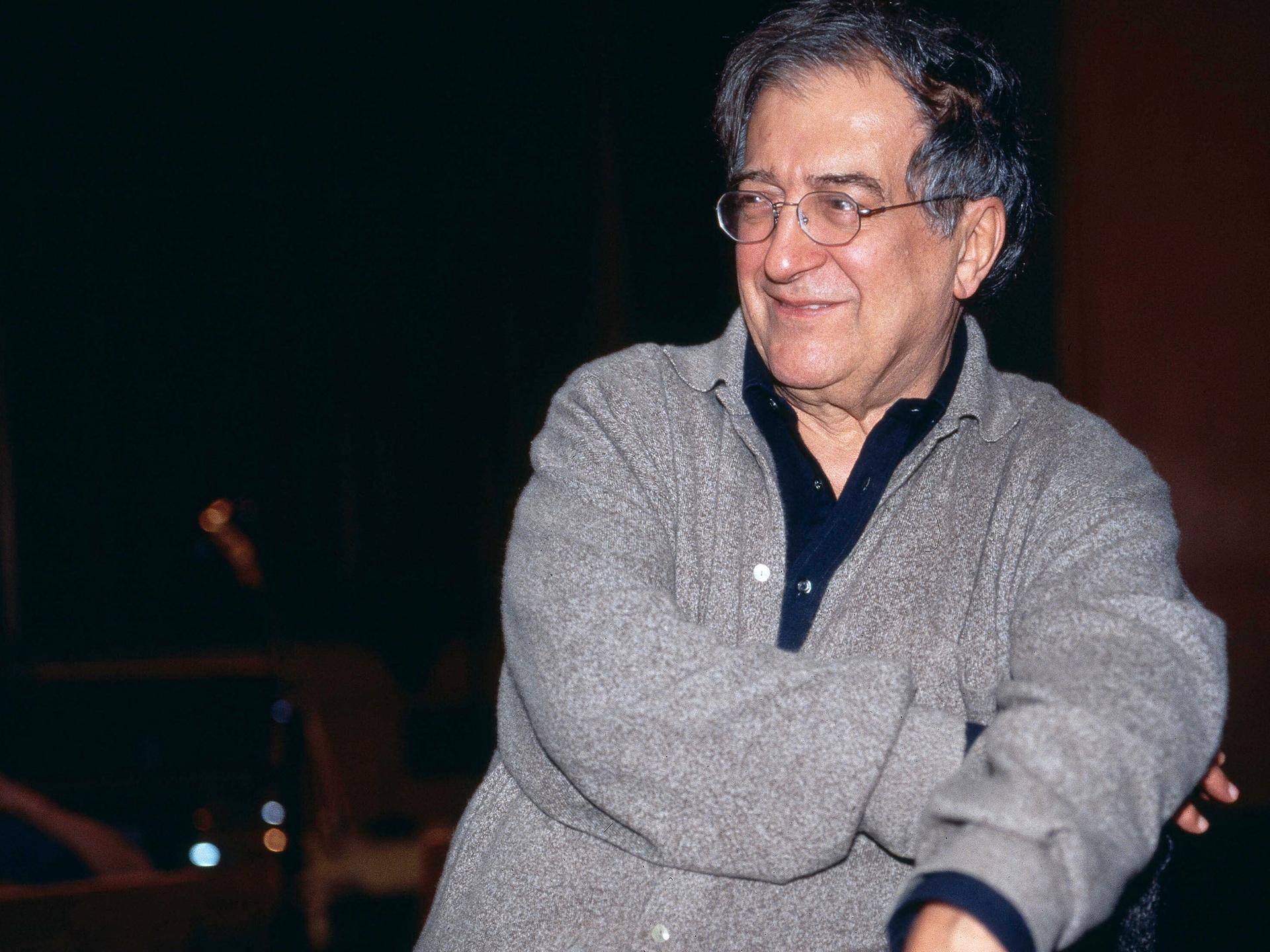 Luciano Berio sitzt in grauer Strickjacke und mit Brille in einer Opernprobe und lacht seitlich an der Kamera vorbei.