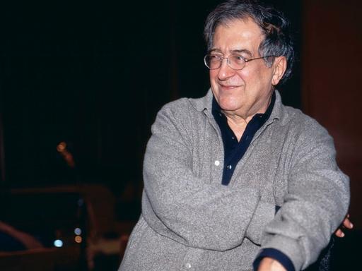 Luciano Berio sitzt in grauer Strickjacke und mit Brille in einer Opernprobe und lacht seitlich an der Kamera vorbei.