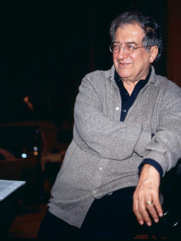 Luciano Berio sitzt in grauer Strickjacke und mit Brille in einer Opernprobe und lacht seitlich an der Kamera vorbei.