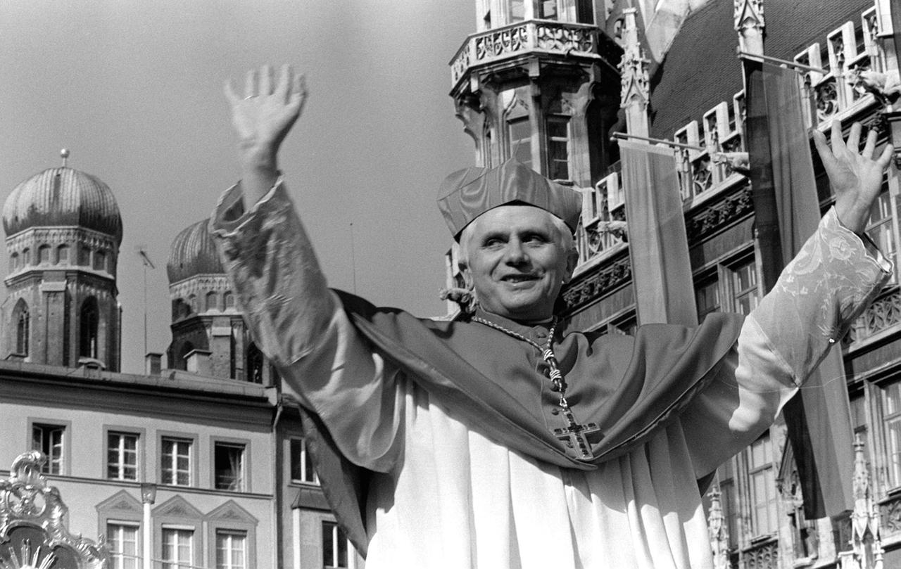 Kardinal Joseph Ratzinger hebt beide Arme zum Gruß, im Hintergrund die Türme des Münchener Doms Liebfrauenkirche