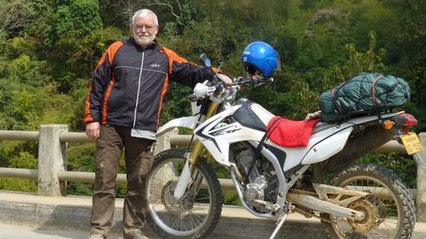 Friedrich Weidner steht neben seinem voll bepackten Motorrad auf einer Straße.