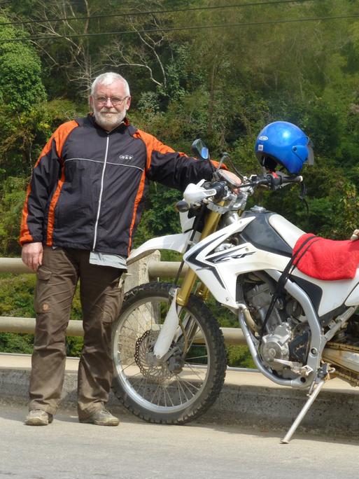 Friedrich Weidner steht neben seinem voll bepackten Motorrad auf einer Straße.