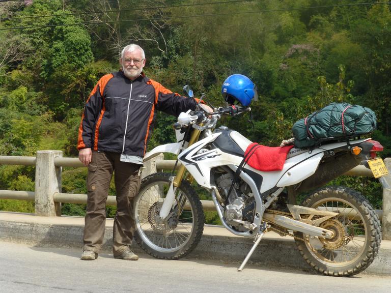 Friedrich Weidner steht neben seinem voll bepackten Motorrad auf einer Straße.