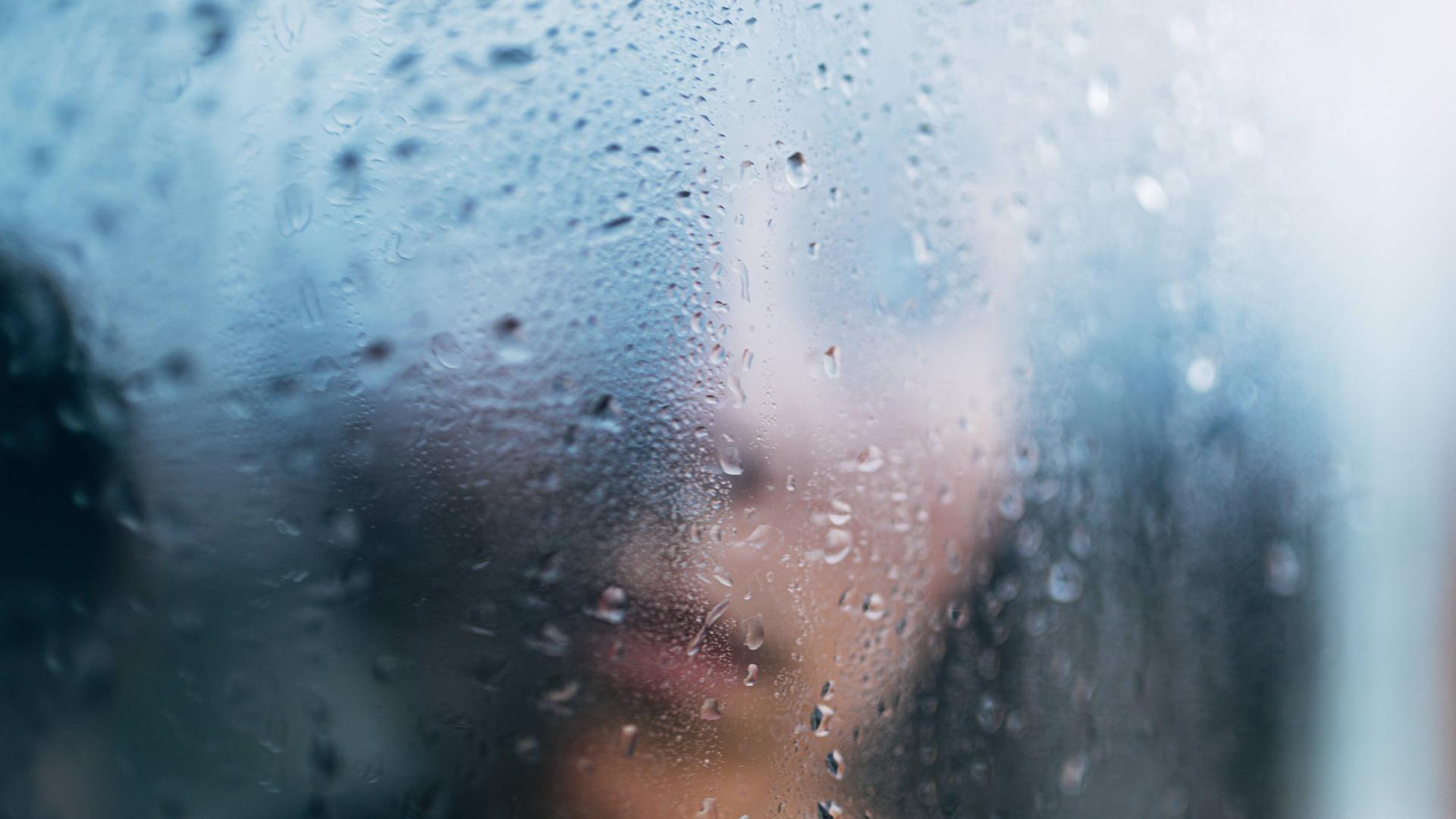 Eine Person steht in Unschärfe hinter einem regennassen Fenster.