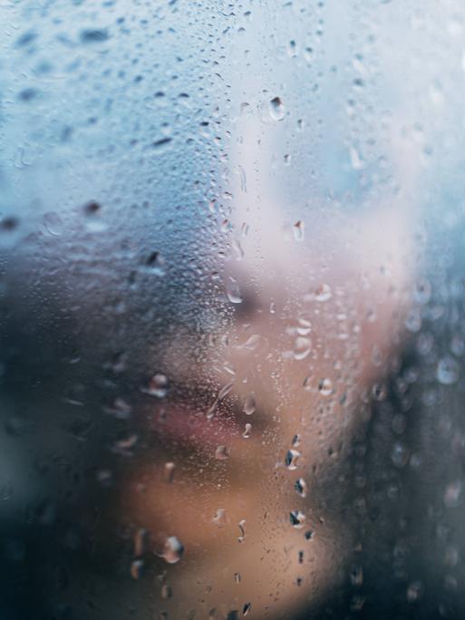 Eine Person steht in Unschärfe hinter einem regennassen Fenster.