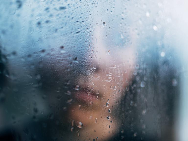 Eine Person steht in Unschärfe hinter einem regennassen Fenster.