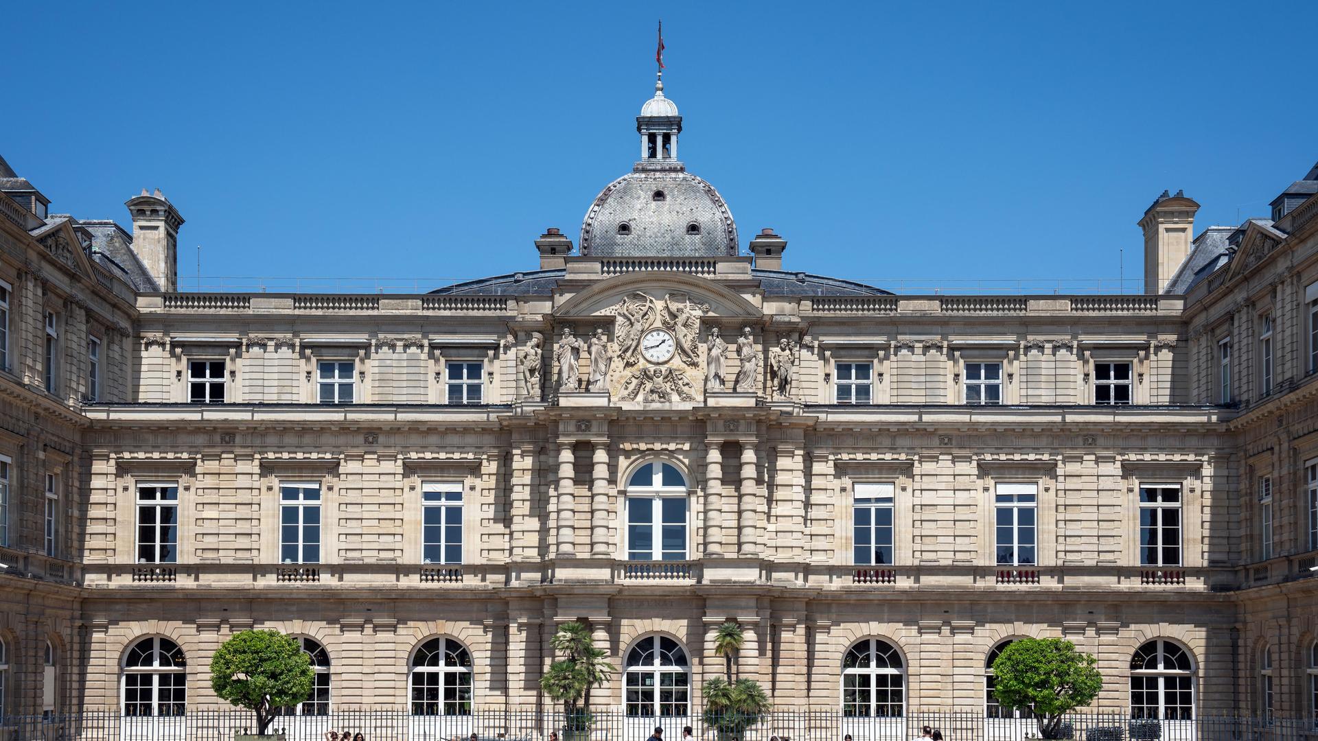 Ein großes Gebäude aus hellem Stein hinter einer Wiese, in der Mitte ragt ein kleiner Turm mit einer Uhr auf.