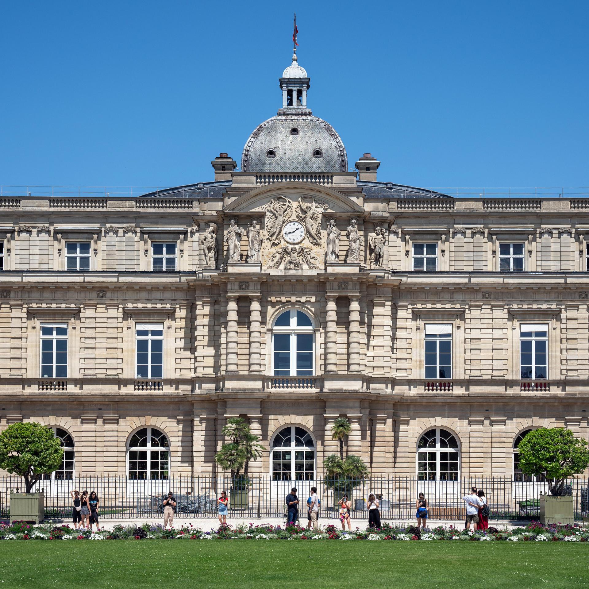 Ein großes Gebäude aus hellem Stein hinter einer Wiese, in der Mitte ragt ein kleiner Turm mit einer Uhr auf.