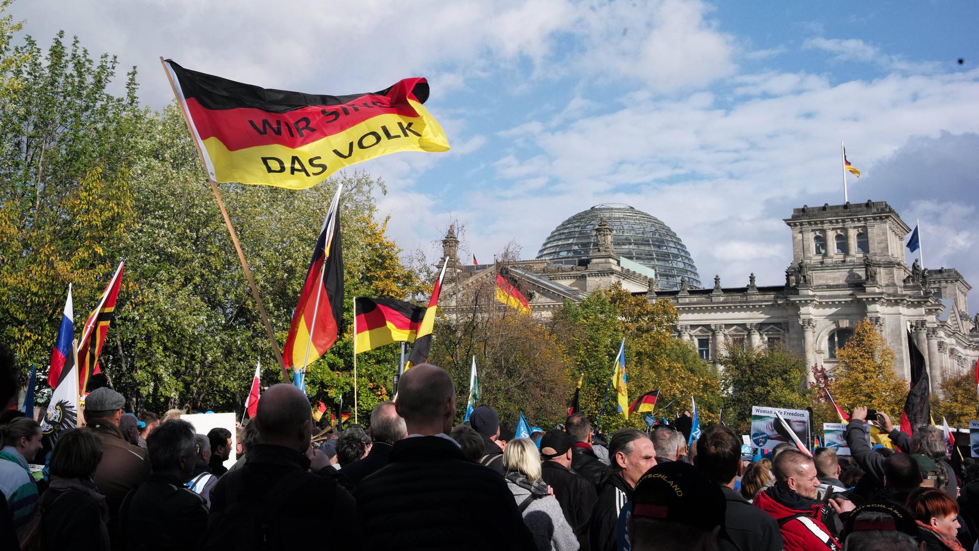 Eine Menschenmenge schwenkt Deutschlandfahnen. Auf einer davon ist zu lesen: "Wir sind das Volk". Im Hintergrund: der Reichstag, Sitz des deutschen Bundestags.