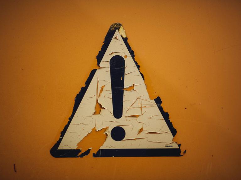 Auf gelbem Hintergrund klebt ein schwarz umrandetes weißes dreieckiges Schild mit einem Ausrufezeichen. 