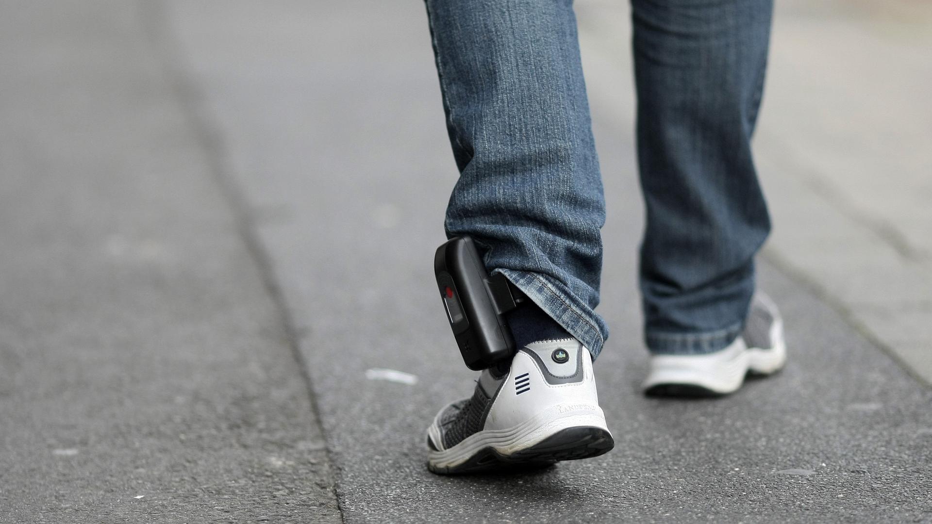 Man sieht die untere Beine eines Mannes mit einer elektronischen Fußfessel über seiner Jeans. 