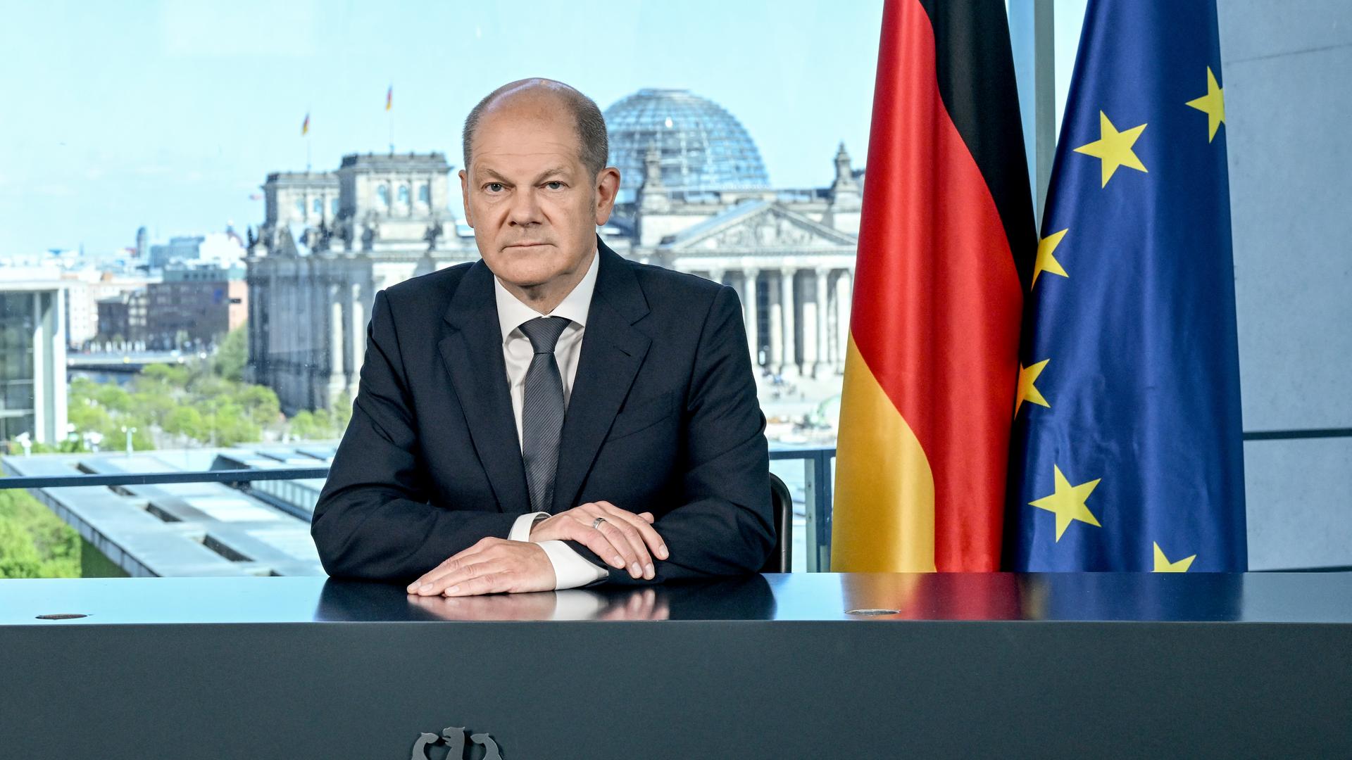 Kanzler Scholz sitzt hinter einem Pult mit Bundesadler. Neben ihm die deutsche Fahne unb die Europaflagge, im Hintergrund ist der Reichstag zu sehen. 